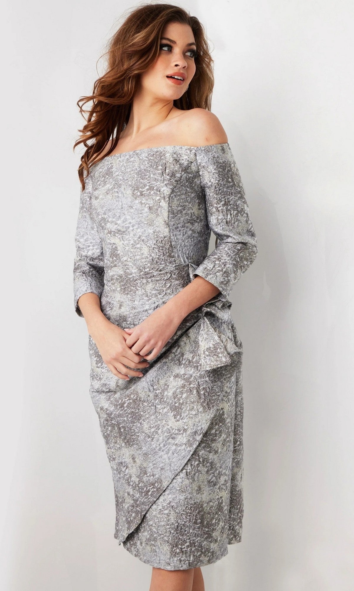A woman wearing the Jovani 09551 short wedding guest dress in silver/grey, featuring an off-the-shoulder neckline, long sleeves, knee-length hem, and textured patterned fabric, stands against a plain background.
